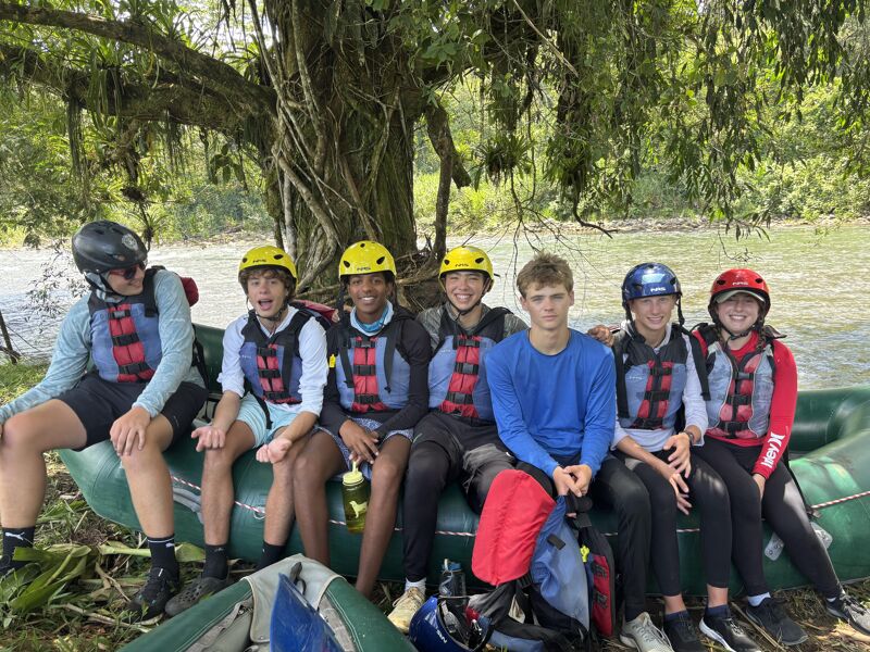 The image shows a group of six young people wearing helmets and life jackets, sitting on the edge of an inflatable raft. They appear to be preparing for or have just finished a rafting trip. The background features a river and lush green vegetation, suggesting a tropical or forested environment. The group is smiling and seems to be enjoying their outdoor adventure.
